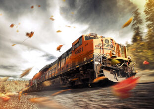A freight train with the number 6991 speeds along a track, surrounded by swirling autumn leaves, against a blurred backdrop of trees and a cloudy sky.