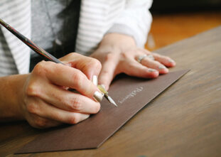 A hand holds a calligraphy pen and is writing script letters in silver on a brown paper