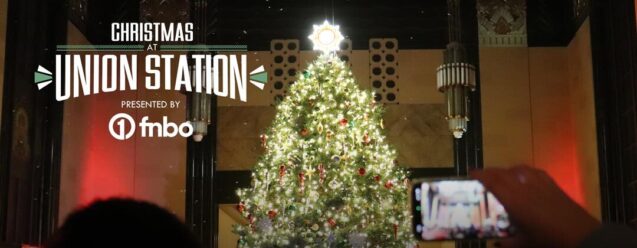 A tall, decorated Christmas tree and choir in a big hall with a “Christmas at Union Station” event graphic.