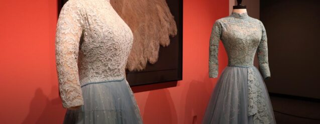 Two blue lace-and-tulle dresses displayed on mannequins beside a large feather fan.
