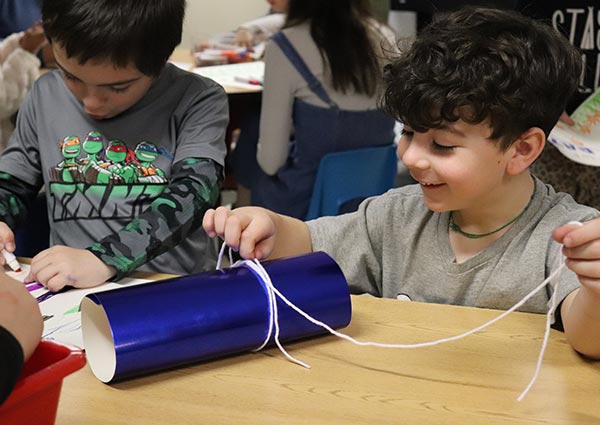 Two children work on a craft project involving a blue cardboard tube and white string at a table.